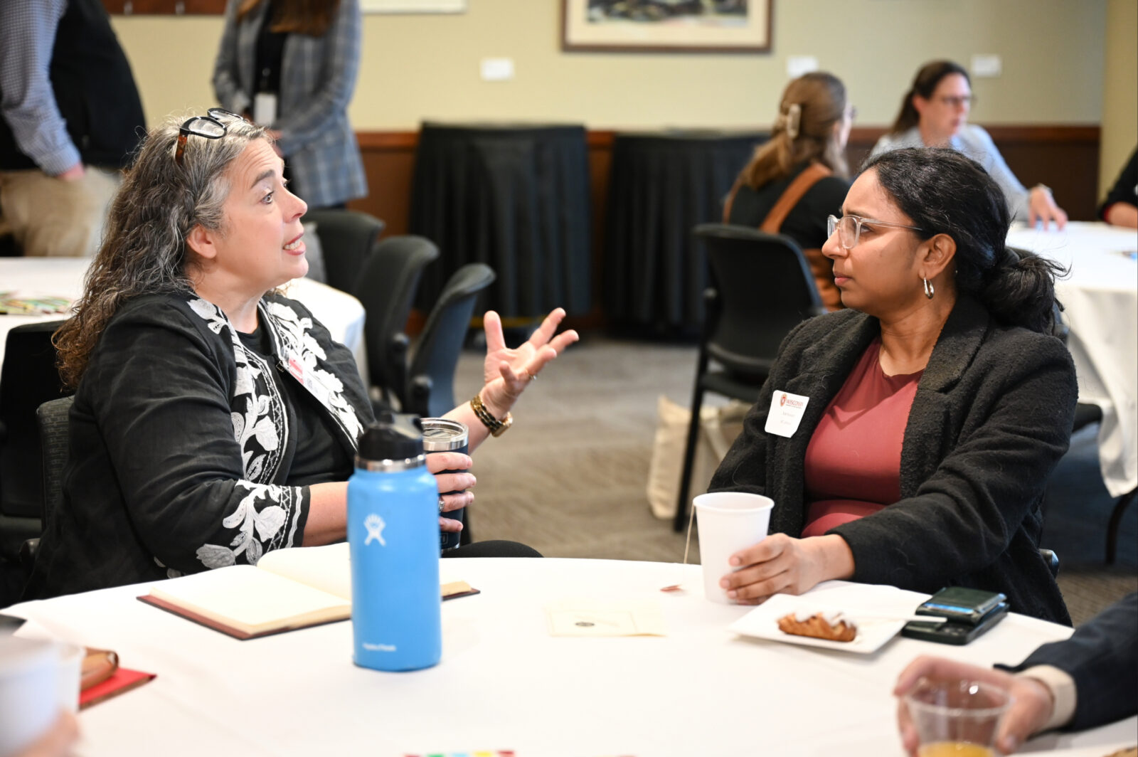 Two individuals are facing and talking to each other, seated at a table. Food and drinks are on the table.
