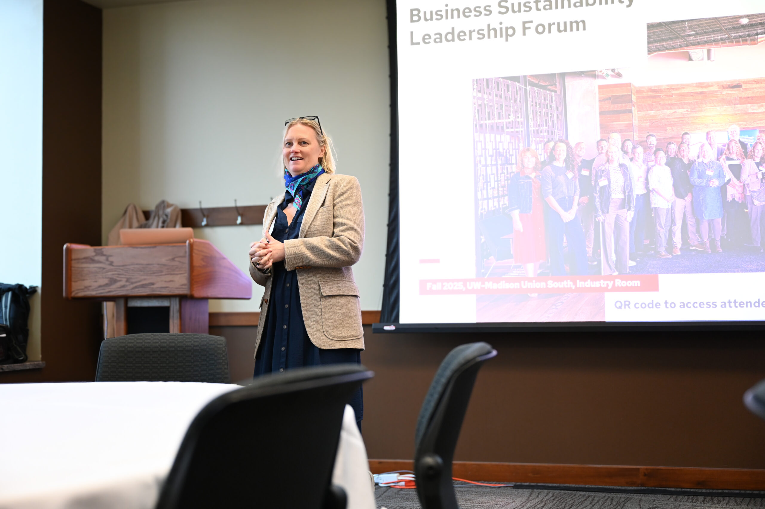 Someone in a blazer is presenting in the front of a room. Behind her is a slide that reads "Business Sustainability Leadership Forum."