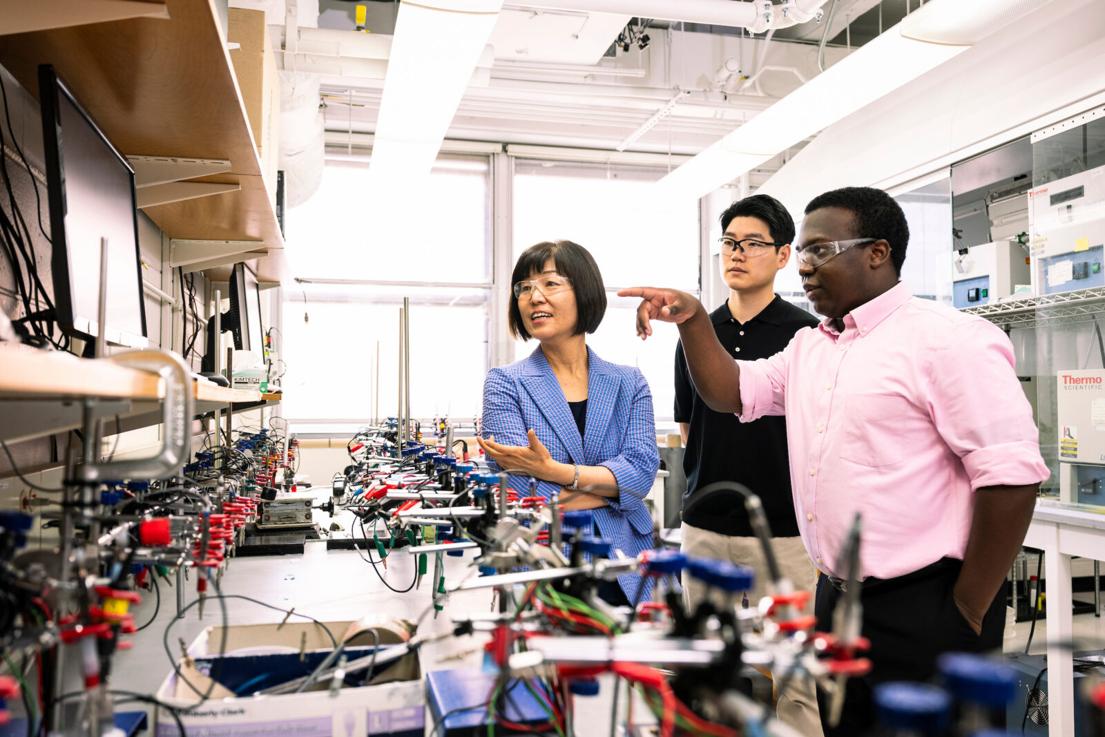 Three people with safety glasses on look at something together in a chemistry lab.