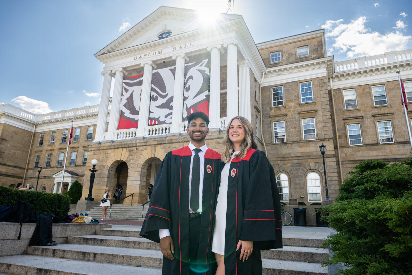 Two students in graduation gowns pose in front of Bascom Hall.