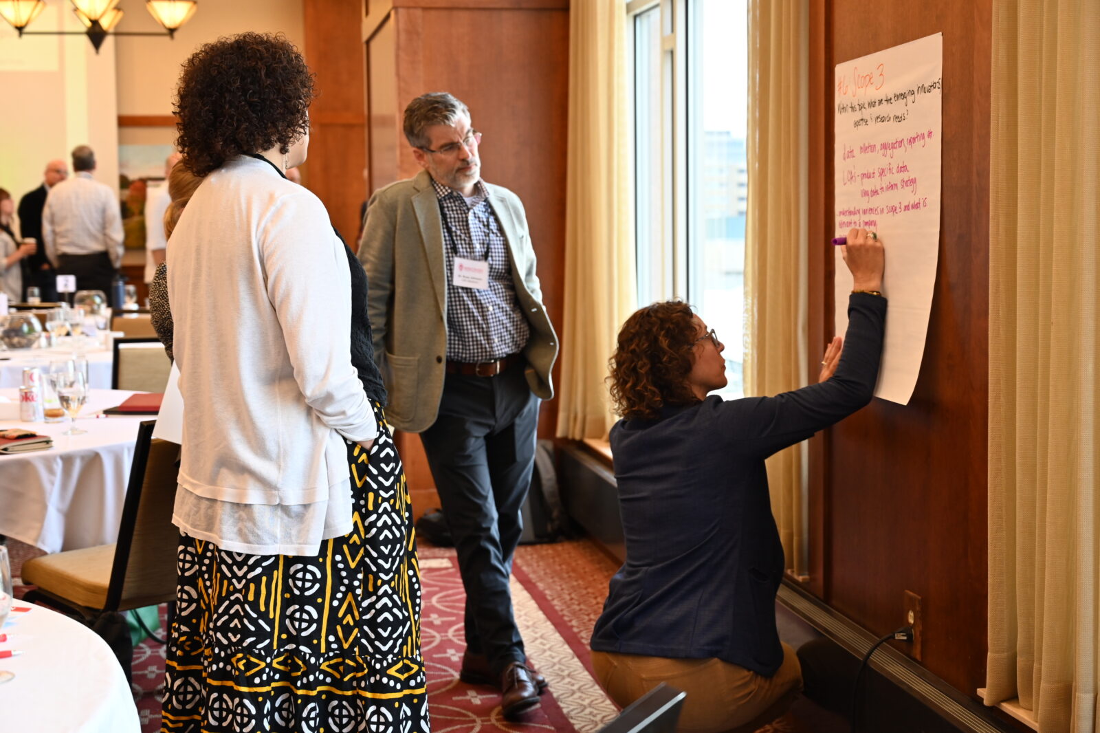 A group of people brainstorms together. One of them takes notes on a large Post-it note on the wall.